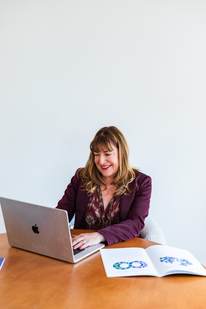 Antoinette Oglethorpe sits at a desk working on a laptop, smiling. She wears a maroon blazer and patterned blouse. An open booklet with colourful graphics is on the table beside her. The background is plain white.