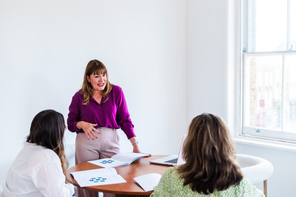 Antoinette Oglethorpe in a purple blouse stands and speaks to two seated people delivering one of her career conversations masterclasses at a round table with documents and a laptop in a bright, white-walled office.