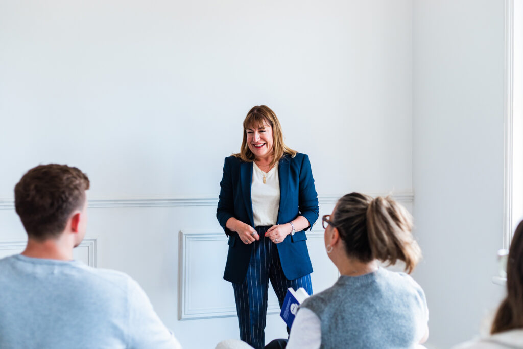 Antoinette Oglethorpe in a blue blazer is smiling and speaking to a group of people seated in a bright, white-walled room enjoying her career management masterclass