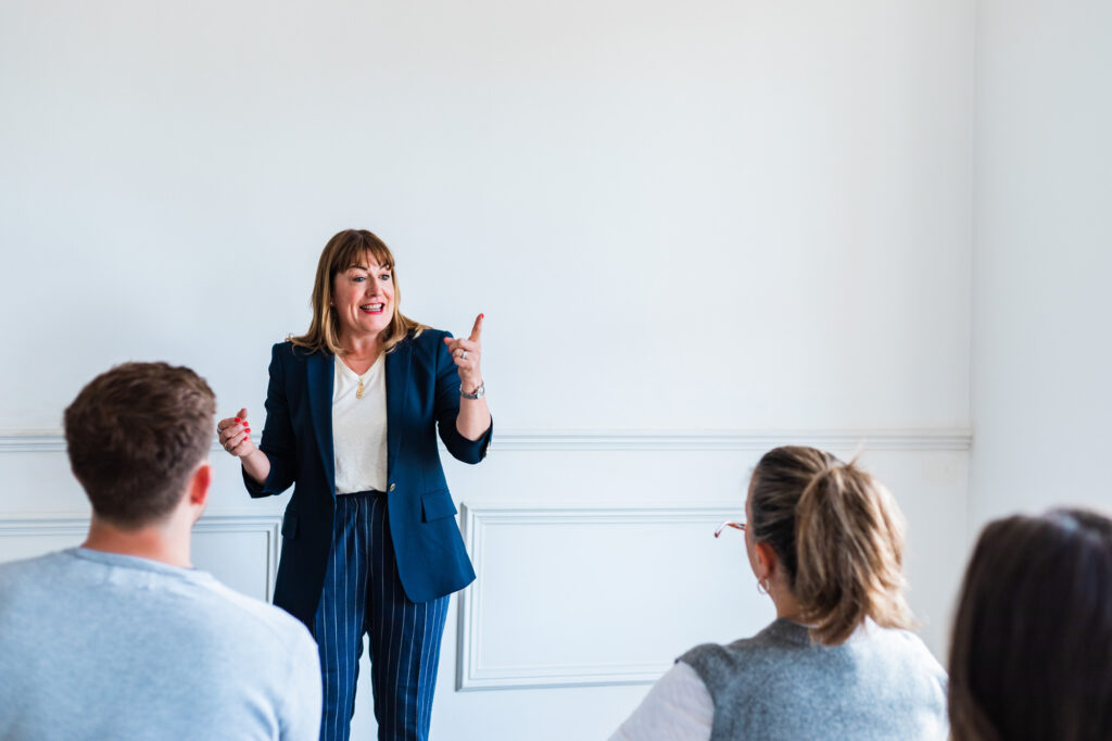 Antoinette Oglethorpe in a jacket stands speaking and gesturing to a group of seated people in a bright room with white walls.