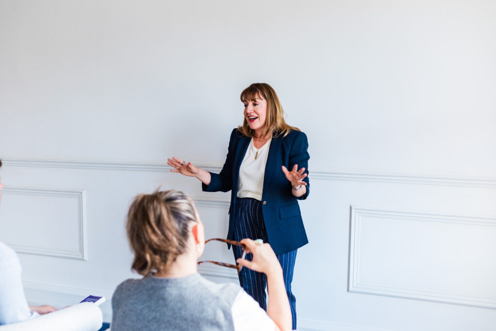 Antoinette Oglethorpe in a blazer stands and speaks enthusiastically to a seated audience, gesturing with her hands in a bright, minimalist room. One member of the audience holds spectacles and listens attentively.