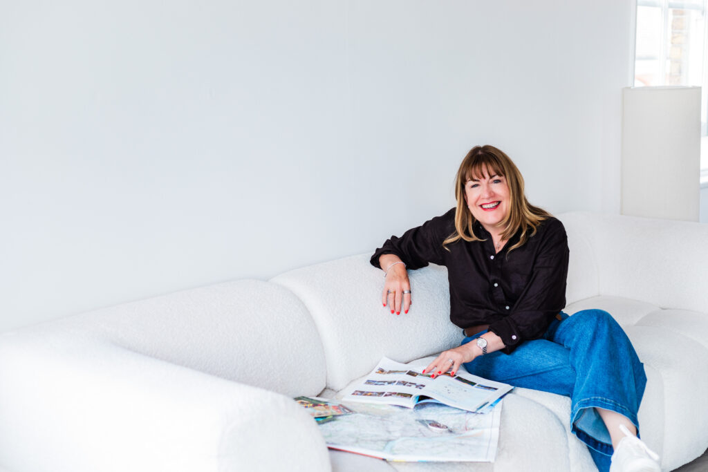 Antoinette Oglethorpe author of the COMPASS Journal blog, wearing a black shirt and blue jeans, sits smiling on a white sofa with magazines and papers spread out in front of her in a bright, minimalist room.