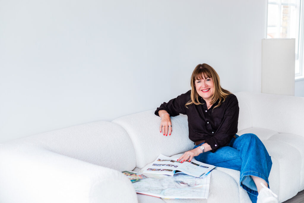 A woman with straight brown hair, wearing a black shirt and blue jeans, sits on a white sofa, smiling and holding open magazines on her lap in a bright, minimalist room.