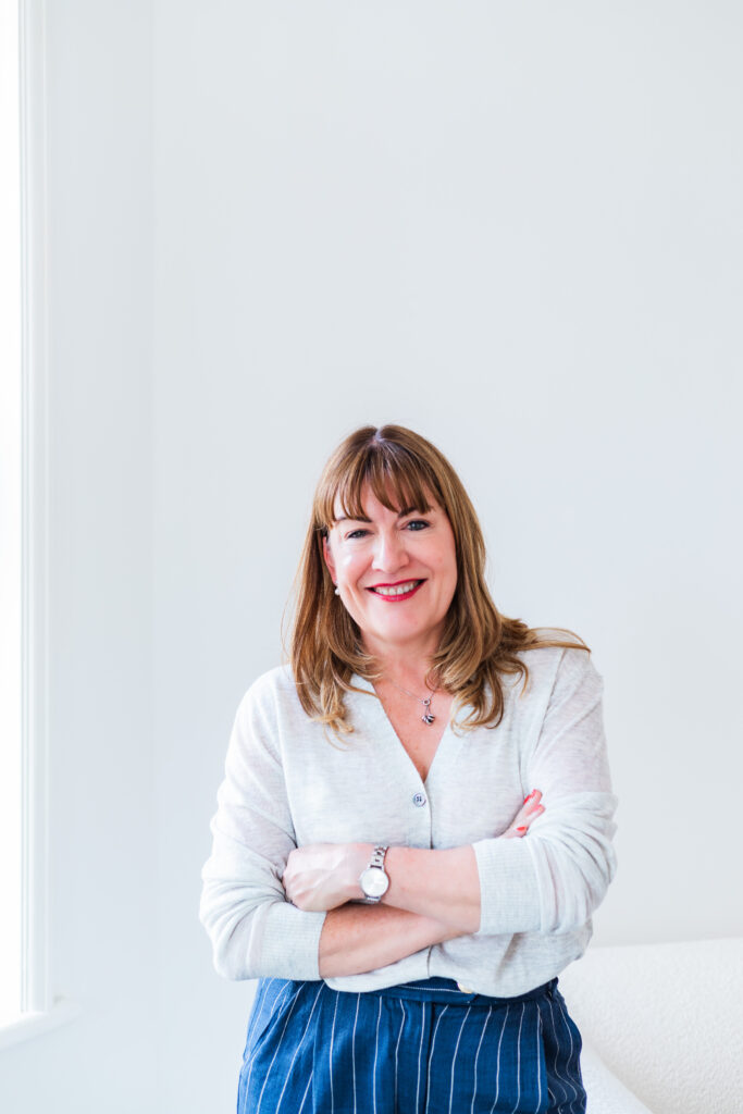 Antoinette Oglethorpe headshot, she has medium-length brown hair and a fringe stands smiling with arms crossed, wearing a light jumper and blue pinstripe trousers, against a plain white background near a window.