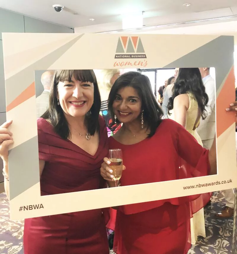 Two women in red dresses smile and pose together, holding a large photo frame labelled "National Business Women's Awards" at a formal event. One woman holds a glass of champagne.