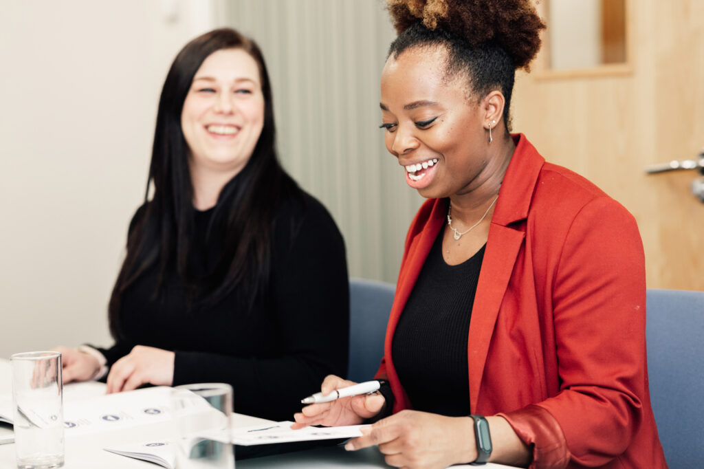 Two women sit at a table, smiling and holding pens while looking at documents. One wears a red blazer and the other wears black. They appear to be in a meeting or collaborative work setting.