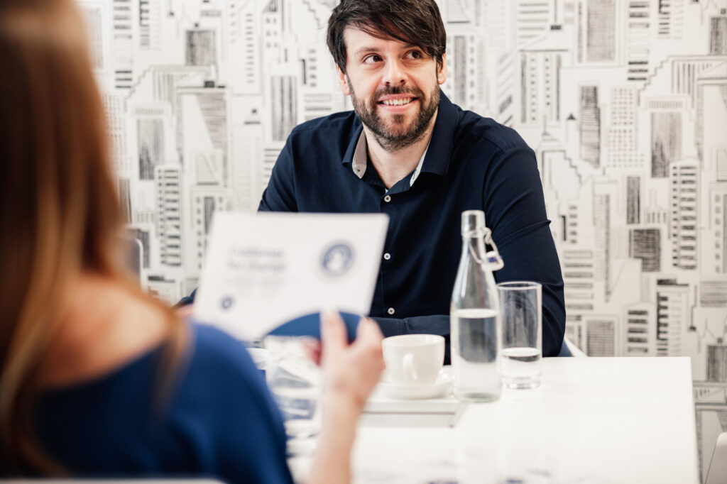 A man with dark hair and a beard smiles whilst sitting at a table with a cup, glass of water, and bottle, talking to a woman holding a document, in a modern office with a cityscape wallpaper background, during a training session for the Certified Facilitator Programme designed and delivered by Antoinette Oglethorpe.
