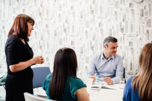 Four people are in a meeting room. Antoinette Oglethorpe stands speaking to three seated colleagues, one man and two women, who are listening and looking at a book on the table. The background has white wallpaper with building designs.