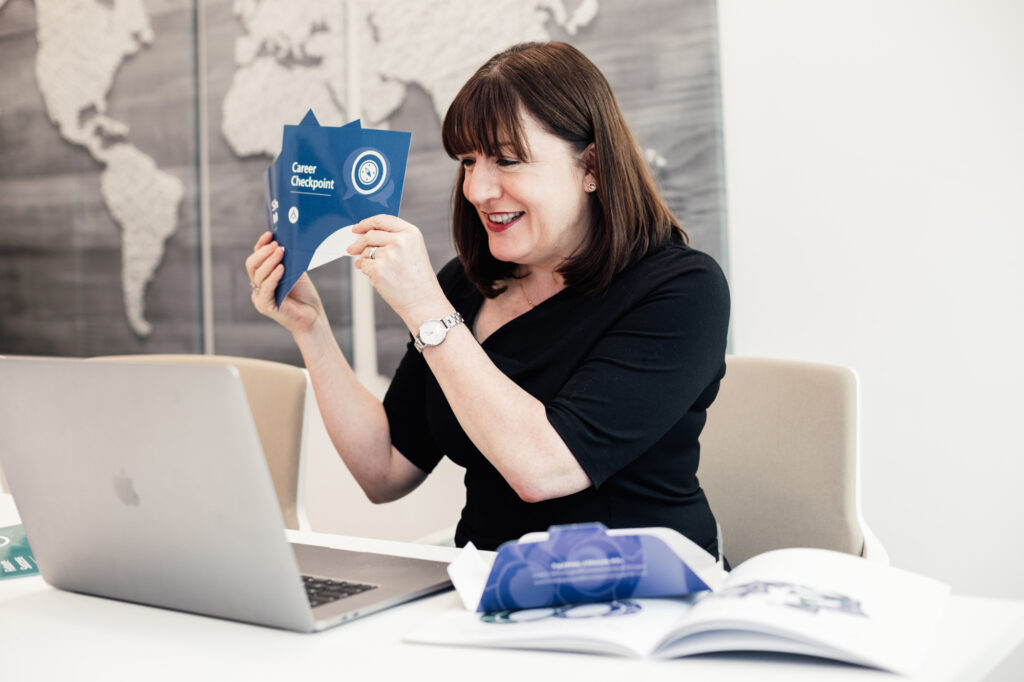 Antoinette Oglethorpe sits at a desk smiling whilst holding up a card from her Career Conversation Toolkit titled 
