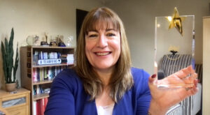 Antoinette Oglethorpe holds up a clear glass award with a gold star in a cosy room with bookshelves, plants, and a 