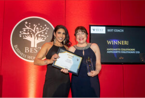 Two women in black dresses smile and hold a certificate and trophy at an awards event. Behind them, a sign reads 