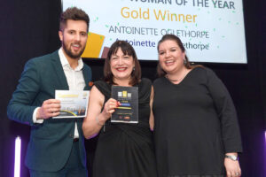 Three people stand together at an awards ceremony, smiling at the camera. Antoinette Oglethorpe is in the centre holds a trophy plaque, while the man on the left holds a 