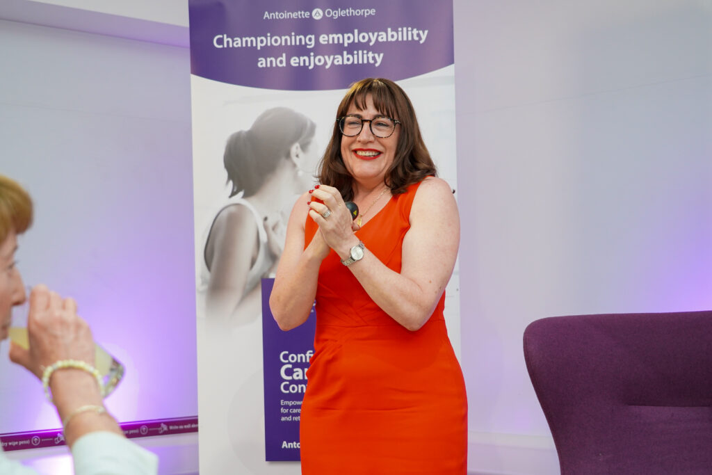 Antoinette Oglethorpe in a bright red dress stands smiling and clapping in front of a banner about employability and enjoyability at an event. A seated person and a purple chair are partially visible in the foreground.