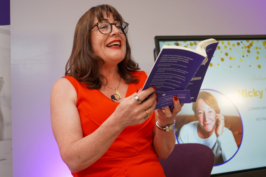 Antoinette Oglethorpe in a bright red dress and glasses smiles whilst reading from her best selling book Confident Career Conversations. Behind her, a screen displays the name 