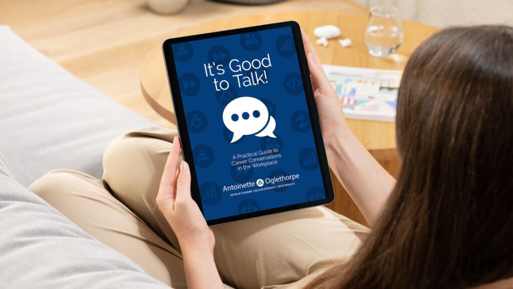 A woman sits on a sofa holding a tablet displaying a blue cover titled “It’s Good to Talk! A Practical Guide to Career Conversations in the Workplace” by Antoinette at Oglethorpe, with a chat bubble icon in the centre.