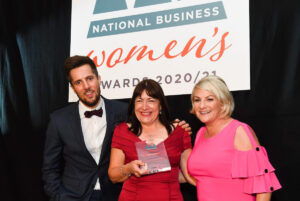 Three people pose and smile at the National Business Women's Awards 2020/21. Antoinette Oglethorpe is in the centre holds an award trophy, with a man in a suit and another woman in a pink dress standing beside her.