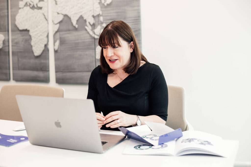 Antoinette Oglethorpe, is sat looking at her laptop smiling and presenting a webinar. Open books and a lanyard are on the desk, and a textured world map artwork hangs on the wall behind her.