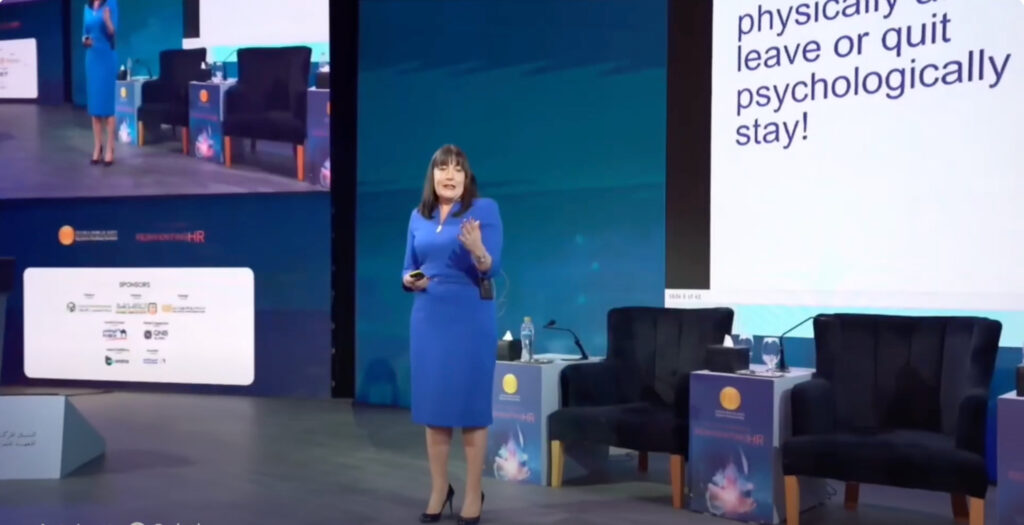 Antoinette Oglethorpe in a blue dress speaks on stage speaking at a conference. Behind her is a large screen with text reading, 