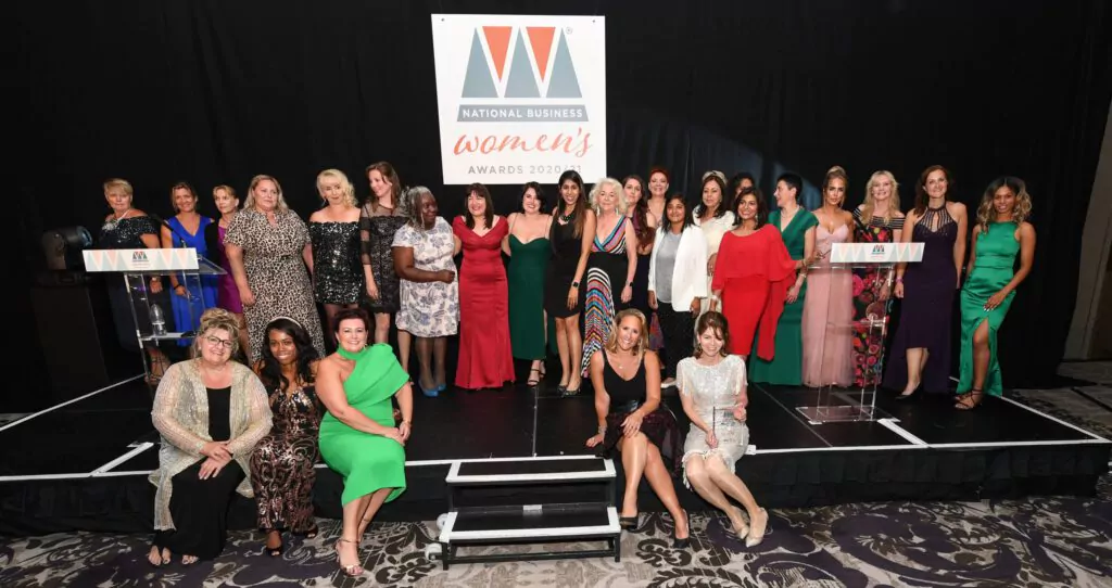 A group of women in formal attire pose together on a stage at the National Business Women’s Awards 2021, with a large event banner behind them and two lecterns on each side of the stage.