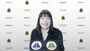 Antoinette Oglethorpe with brown hair and a fringe, wearing a black polka dot top, smiles at the camera. She holds two Stevie Awards. The background displays sponsor logos and 