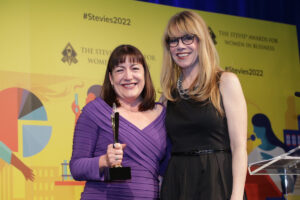 Antoinette Oglethorpe and the award host are smiling at an awards ceremony; Antoinette holds a trophy. They stand in front of a yellow backdrop with the text “#Stevies2022” and “The Stevie Awards for Women in Business”.