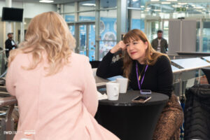 Two women sit at a round table in a modern indoor setting, engaged in conversation. Antoinette Oglethorpe is smiling and listening attentively, with two coffee mugs and a mobile phone on the table—capturing the spirit of IWD and meaningful connection.