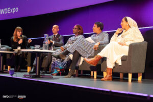 Five panellists sit on stage in conversation at a conference celebrating IWD. Four women of different ethnicities and ages, including Antoinette Oglethorpe who is hosting the panel, and one man are seated on sofas, with microphones and water glasses in front of them. A purple backdrop displays event branding.