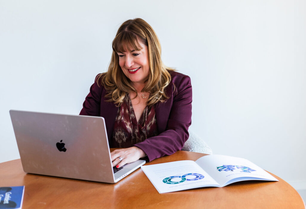Antoinette Oglethorpe with brown hair smiles whilst working on a laptop at a wooden table. Delivering the Certified Facilitator Programme a blended live online programme and self paced course work for HR leaders An open booklet with colourful charts or diagrams lies next to her on the table. The background is plain and white.