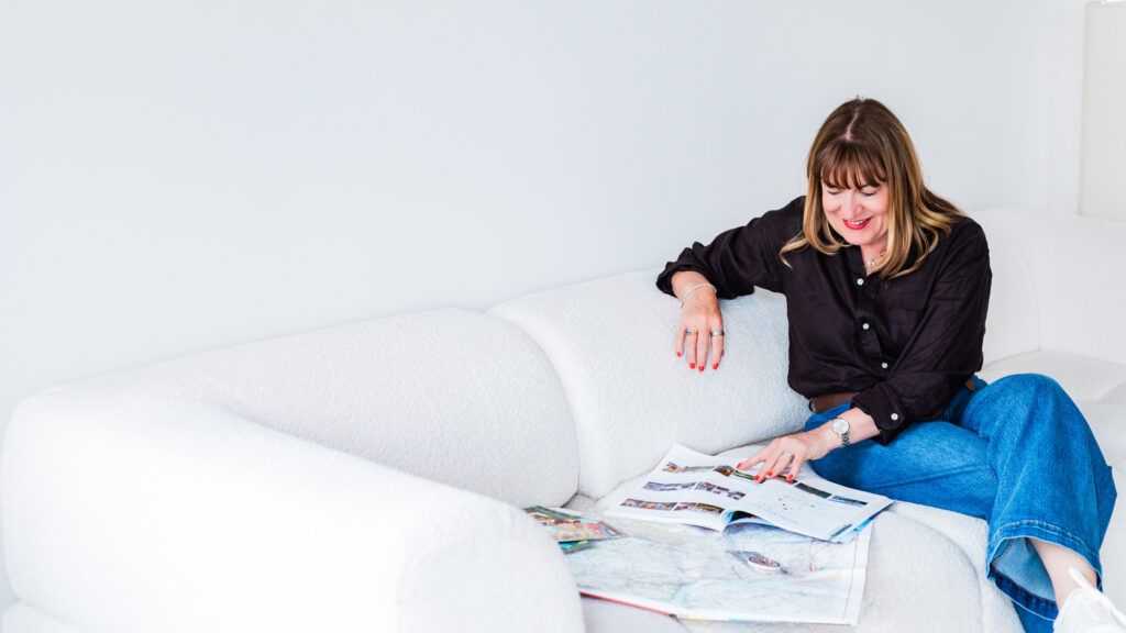 Antoinette Oglethorpe has straight brown hair, wearing a black shirt and blue jeans, sits on a white sofa and smiles while looking through magazines spread on the seat beside her.