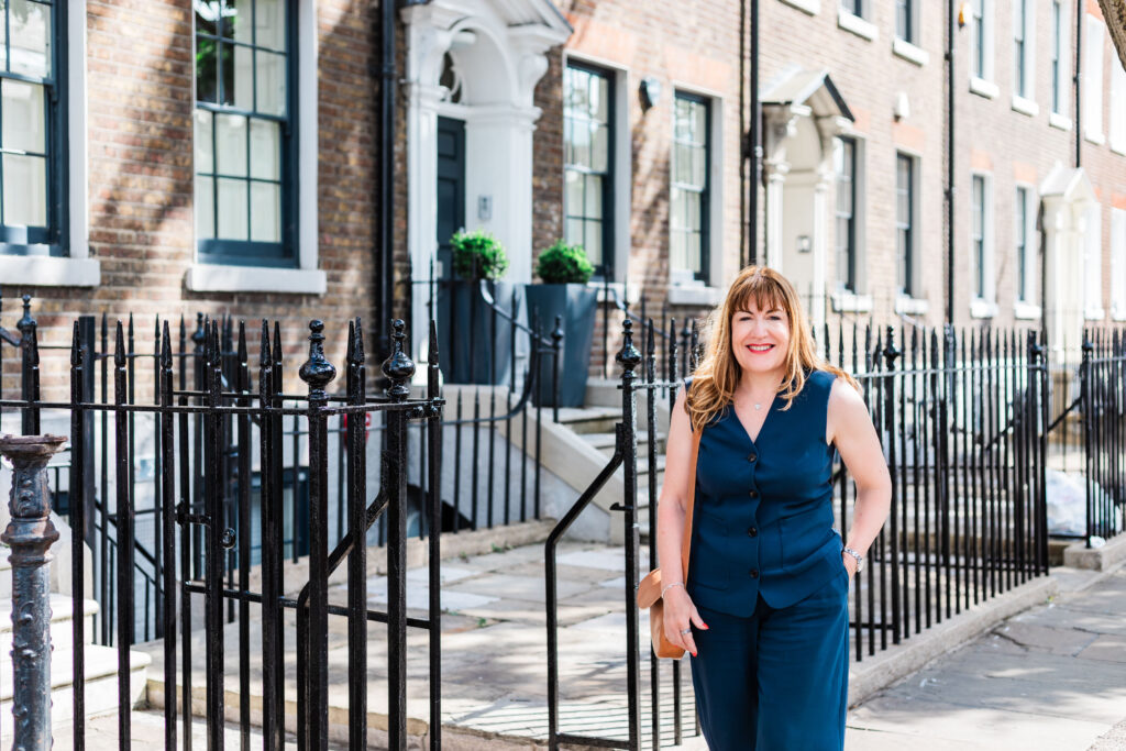 Antoinette Oglethorpe with light brown hair wearing a navy blue sleeveless outfit stands smiling on a sunny pavement in front of Georgian-style terraced houses with black railings.