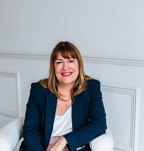 Antoinette Oglethorpe headshot, she has light brown hair and fringe, wearing a dark blazer and white top, smiles while sitting in a white chair against a light-coloured wall with decorative moulding.