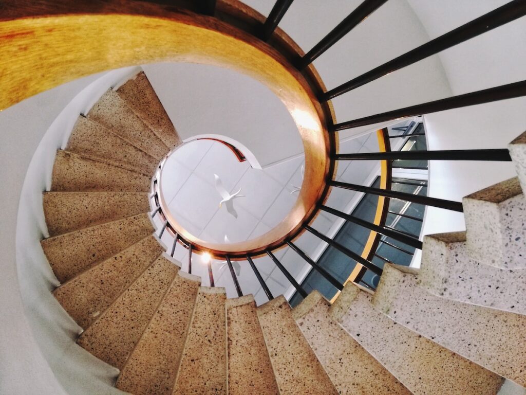 A spiral staircase with beige steps and black railings, viewed from above. The curved handrail is light brown, and the stairwell forms a circular pattern—resembling career progression—leading down to a white floor and ceiling fan.