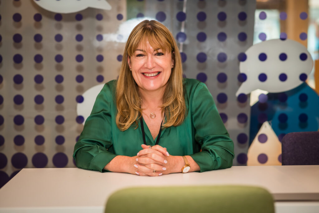 Antoinette Oglethorpe shares her exceptional client experiencxe stories and case studies, image of Antoinette with long honey blonde hair, wearing a green blouse, sits at a white table smiling. The background features a glass wall with purple dots and white speech bubble shapes.
