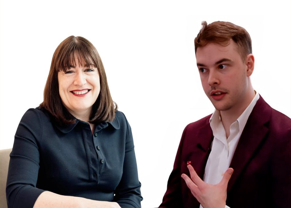 Two people against a white background: a woman with straight dark hair in a black top smiling, and a man in a maroon jacket and white shirt gesturing with one hand whilst speaking.