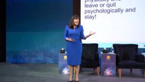 Leading career management and leadership speaker Antoinette Oglethorpe is on stage as the keynote speaker in a blue dress speaks on large stage in front of a screen displaying text.