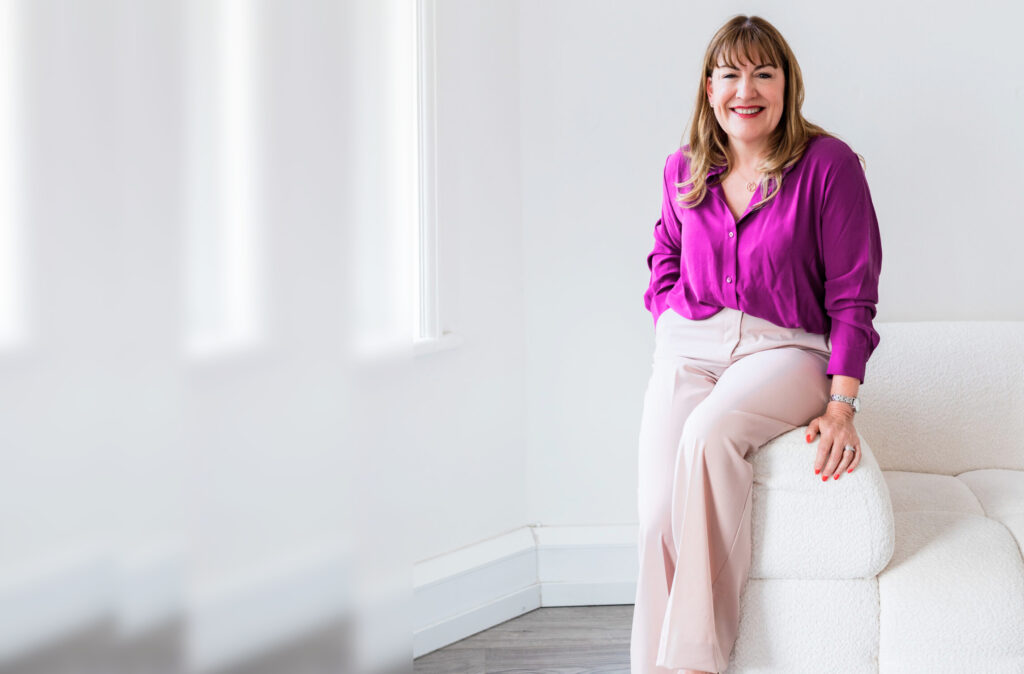 Antoinette Oglethorpe wearing a bright purple blouse and light pink trousers, sits smiling on a modern white sofa in a bright, minimalist room with large windows.