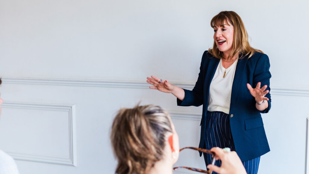 Antoinette Oglethorpe wearing a navy blazer and striped trousers stands in front of a white wall, smiling and gesturing with her hands, whilst speaking to two seated people, one holding spectacles.