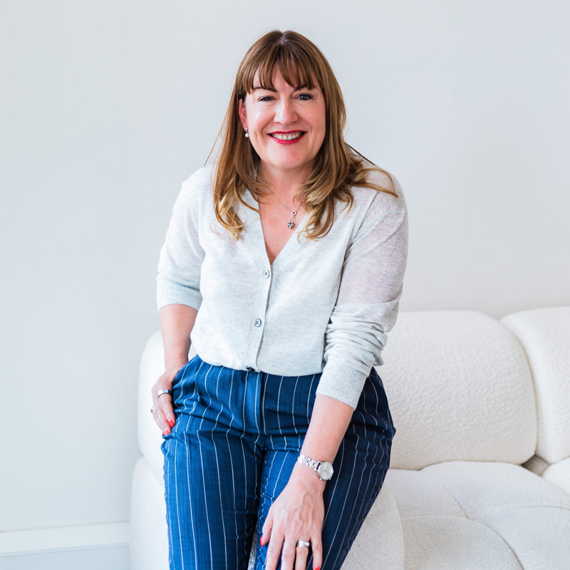 Antoinette Oglethorpe profile picture wearing a light cardigan and blue pinstripe trousers, smiles while sitting on a white sofa against a plain light background.