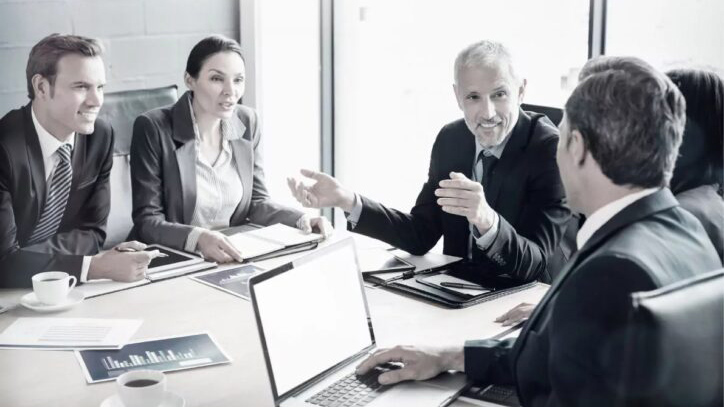 Five business professionals in suits have a serious discussion round a conference table with laptops, papers, and charts in a bright office setting. One man gestures whilst others listen and take notes.