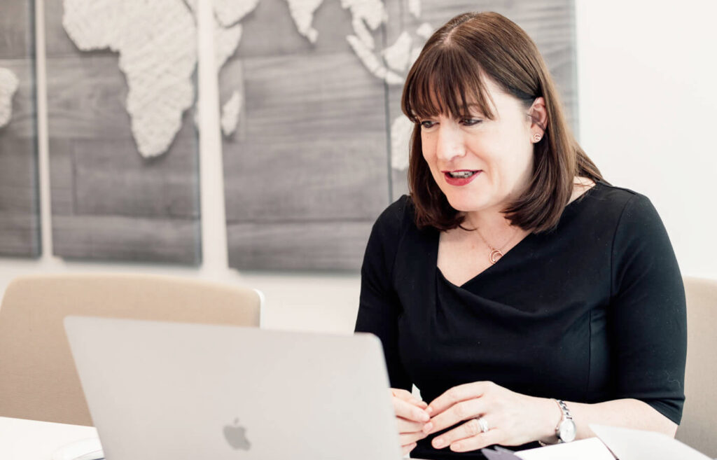 Antoinette Oglethorpe delivers the confident career conversations webinar wearing a black top, sits at a table and presents on a laptop. A world map wall art is visible in the background.
