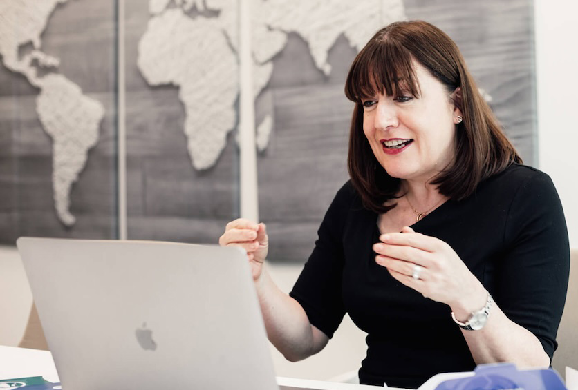A woman with shoulder-length brown hair sits at a desk, smiling and gesturing with her hands whilst looking at a laptop. A world map artwork is visible on the wall behind her.