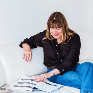 Antoinette Oglethorpe has straight brown hair, wearing a black shirt and blue jeans, sits on a white sofa and smiles while looking through magazines spread on the seat beside her.