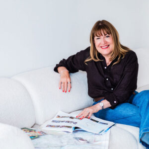 A woman with straight brown hair, wearing a black shirt and blue jeans, sits on a white sofa, smiling and holding open magazines on her lap in a bright, minimalist room.