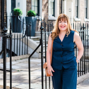 Antoinette Oglethorpe with light brown hair wearing a navy blue sleeveless outfit stands smiling on a sunny pavement in front of Georgian-style terraced houses with black railings.