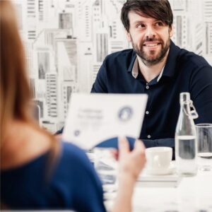 A man with dark hair and a beard smiles whilst sitting at a table with a cup, glass of water, and bottle, talking to a woman holding a document, in a modern office with a cityscape wallpaper background, during a training session for the Certified Facilitator Programme designed and delivered by Antoinette Oglethorpe.