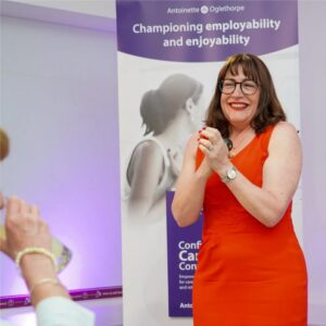 Antoinette Oglethorpe in a bright red dress stands smiling and clapping in front of a banner about employability and enjoyability at an event. A seated person and a purple chair are partially visible in the foreground.