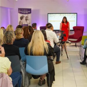 Antoinette Oglethorpe stands and speaks in front of an audience seated in a modern, brightly lit room with colourful chairs, a large screen, and posters on the walls at her book launch party celebrating her Amazon #1 best seller Confident Career Conversations.
