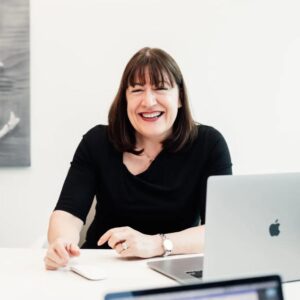 Antoinette Oglethorpe is in a black top and is smiling whilst sitting at a desk with an open laptop. A map is on the wall behind her, she has just delivered the webinar how to design and implement a mentoring programme that maintains momentum